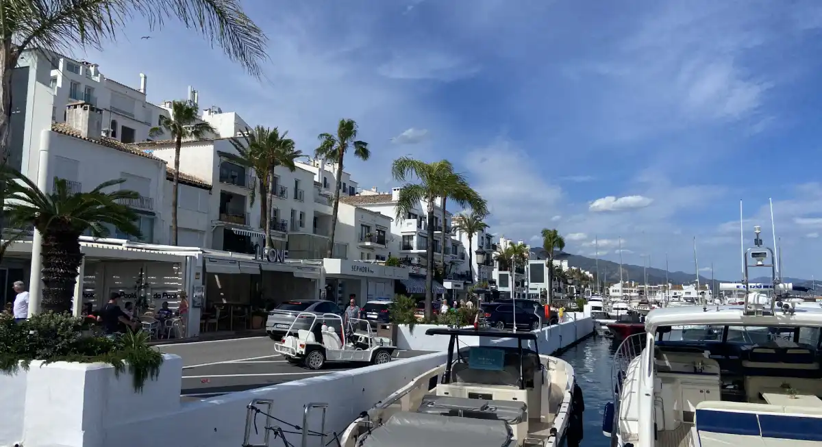 Puerto Banús marina with luxury yachts, white Mediterranean buildings, palm trees, and parked cars along the waterfront promenade – ideal car rental pickup location