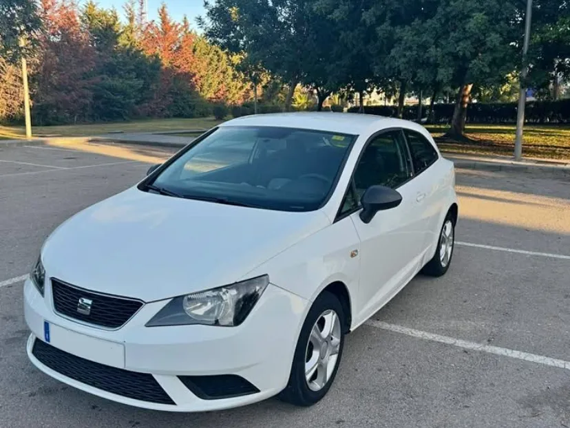 A white SEAT Ibiza is parked in an open, sunlit parking area bordered by tall trees and soft afternoon shadows. The compact hatchback's clean, modern design is highlighted by its sharp headlights, smooth body lines, and well-kept exterior, making it appear practical and reliable for everyday driving. The calm outdoor setting enhances the vehicle's presentation and supports content related to renting cars on the Costa del Sol, making it an ideal visual for travellers searching to rent a car Marbella or rent a SEAT Ibiza Marbella for exploring the region's beaches, city centres, and scenic routes.