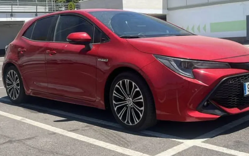 A red Toyota Corolla Hybrid parked in a sunny outdoor lot in Marbella, highlighting its modern hatchback design, aerodynamic front grille, and stylish alloy wheels. This clean and efficient car is ideal for travelers searching to rent a Toyota Corolla in Marbella, offering reliability, comfort, and excellent fuel economy for exploring the Costa del Sol. The bright setting and well-maintained vehicle visually reinforce the convenience and value of choosing this model when looking to rent a car in Marbella.